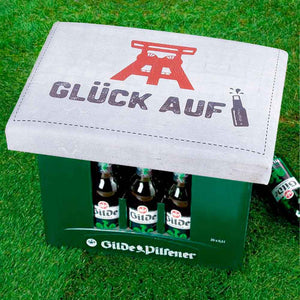 Auf einem Bierkasten ist eine Bierkastensitz mit einer Zeche und dem Spruch: Glück auf zu sehen.