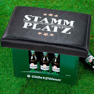 Ein Bierkasten mit einem bequemen Sitzpolster und der Aufschrift Stammplatz steht auf dem Rasen.