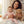 Laden Sie das Bild in den Galerie-Viewer, Das Bild zeigt eine lächelnde Frau mit dunklen Locken und ein Baby, das in ihrem Arm sitzt. Beide schauen in die Kamera. Die Frau hat ein helles, cremefarbenes Oberteil an und hält mit ihrer linken Hand ein braunes Stofftier in Form eines Hundes. Das Baby trägt einen rosafarbenen Strampler und lacht fröhlich. Über den Beinen der Frau liegt eine hellgraue Decke mit weißen Wolken. Der Hintergrund ist ein Wohnzimmer mit einem hellen Teppich.