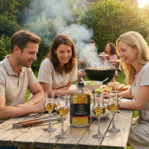 Drei fröhliche Erwachsene, ein Mann und zwei Frauen, sitzen an einem hölzernen Gartentisch. Auf dem Tisch stehen eine Flasche "DIE BERGISCHE MARILLE" und drei gefüllte Schnapsgläser. Im Hintergrund wird auf einem Grill Essen zubereitet, Rauch steigt auf, und weitere Personen sind unscharf zu sehen. Die Szene ist sommerlich und gesellig.