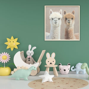 Liebevolle Babyzimmer Wandgestaltung mit dem Gemälde Das quadratische Alpaka-Wandbild hängt an einer salbeigrünen Wand über pastellfarbenem Kinderspielzeug, Dinosauriern und einem Hasen-Holzstuhl im skandinavischen Design.