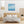 Eine helle Raumansicht einer modern eingerichteten Ferienwohnung, die das quadratische, maritime Acrylgemälde als Wanddekoration über dem Bett präsentiert. Das Gemälde zeigt Segelboote auf dem Meer im Sonnenschein.
