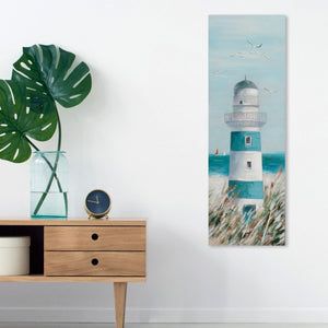 Ein schmaler, vertikaler Kunstdruck eines blau-weißen Leuchtturms hängt an einer schlichten weißen Wand über einem Sideboard aus hellem Holz. Das Gemälde zeigt den Leuchtturm inmitten von hohen Gräsern unter einem hellblauen Himmel mit fliegenden Möwen. Links auf dem Sideboard stehen eine Glasvase mit großen grünen Monstera-Blättern, eine kleine dunkelblaue Uhr und eine weiße Aufbewahrungsbox, was dem Raum eine moderne und maritime Atmosphäre verleiht.