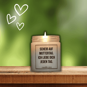 Auf einem Holzbrett im garten steht eine brennende Duftkerze mit dem Spruch: Scheiß auf Muttertag. ich liebe Dich jeden Tag.