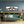 Gerahmtes Panorama-Gemälde mit fünf lustigen Kühen mit Sonnenbrillen und rosa Kunsthaar an einer graublauen Wand über einer Couch.