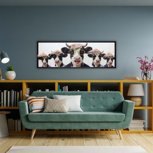 Gerahmtes Panorama-Gemälde mit fünf lustigen Kühen mit Sonnenbrillen und rosa Kunsthaar an einer graublauen Wand über einer Couch.