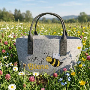 In einer Sommerwiese mit vielen Blumen steht eine hellgraue Filztasche auf der Fleißige Biene steht. Daneben ist eine große Biene eingestickt