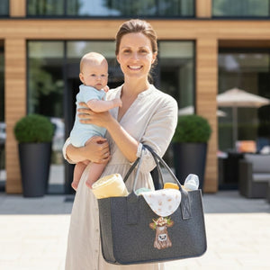 Eine junge Mutter hält ein baby im Arm. Auf der anderen Seite hat sie eine Filztasche mit Babysachen. Auf der Filztasche ist eine Kuh mit buntem Kopfschmuck eingestickt