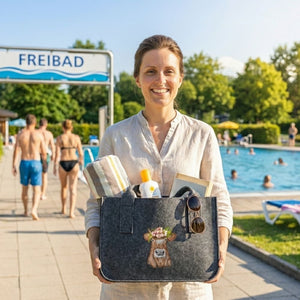 In einem Freibad steht eine Frau mit einer Filztasche in der Badesachen sind. Auf der Filztasche ist eine braune Kuh mit buntem Almkranz eingestickt.