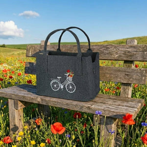 Auf einer Holzbank steht eine Filztasche mit einem eingestickten Fahrrad in der Sonne. Die Szene spielt auf einer bunten Blumenwiese