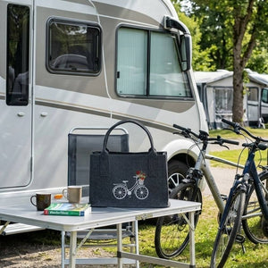 Vor einem Wohnmobil steht eine Filztasche mit einem Fahrrad darauf auf einem Campingtisch. In der Filztasche befinden sich die Lieblingsbücher des Campers.