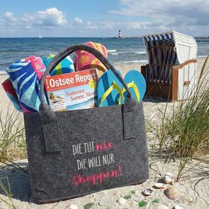 An einem Strand der Ostsee steht ein Filzshopper als Badetasche umfunktioniert. Auf der Filztasche steht der Spruch "Die tut nix, die will nur shoppen"