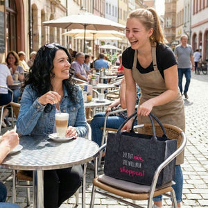 In einer Fußgängerzone befindet sich eine Eisdiele. An einem Tisch draußen sitzt eine Frau mit einem Latte Macciato. Die Bedienung bestaunt ihre Filztasche auf der steht "Die tut nix, die will nur shoppen.