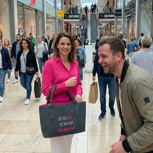 In einem Einkaufcenter steht eine Frau mit einer pinkfarbenen Bluse und hat einen Filzshopper über dem Arm. Auf der Filztasche steht "Die tut nix, die will nur shoppen. Ein Mann steht ihr gegenüber und lacht.