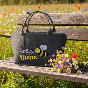 Auf einer Holzbank in einer Blumenwiese steht eine Fliztasche. Auf der Filztasche steht "Fleißige Biene" und eine Biene ist eingestickt. Neben der Tasche liegt ein bunter Blumenstrauß.