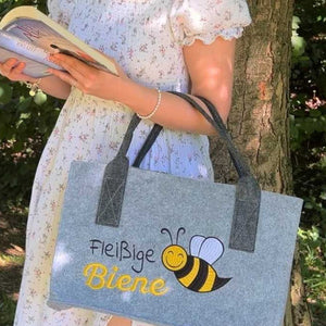 Eine Frau lehnt sich an einen Baum und ließt ein Buch. Über dem Arm trägt sie eine Filztasche mit der Aufschrift "Fleißige Biene"