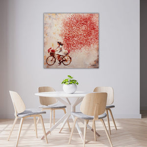 Ein modernes Esszimmer mit Holzstühlen. An der grauen Wand hängt das Bild Flowers in the Air. Es zeigt eine junge Frau auf einem Fahrrad. Vorne am rad hängt eine Blumenkorb der mit weißen und roten Rosen bestückt ist. Hinter dem Fahrrad entsteht eine Wolke aus roten Blumen.