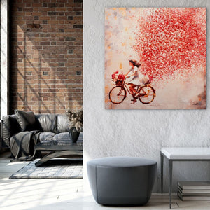 An einer weißen Wand in einem Wohnzimmer hängt ein Gemälde mit einem Mädchen auf dem Fahrrad. Flowers in the Air heißt das Bild. Hinter dem Mädchen ist eine rote Wolke aus Blütenblättern