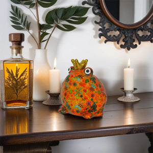 Ein oranger frosch mit dem namen Freddy trägt eine goldene Krone. Der Frosch sitzt auf einem Tisch in einer Wohnung und ist ein wunderschönes Dekoelement.