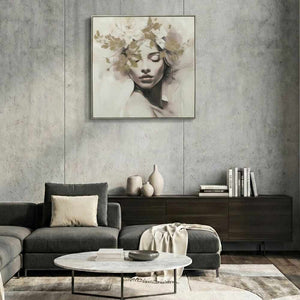 Ein quadratisches Gemälde hängt zentriert über einem grauen Ecksofa an einer Betonwand. Das Kunstwerk zeigt das Porträt einer Frau mit geschlossenen Augen, deren Gesicht von goldenen Blättern und Blumen in Aquarelloptik umrahmt ist. Das Bild ist in einem dünnen, hellen Rahmen eingefasst. Darunter befinden sich ein Marmor-Couchtisch mit Büchern und ein dunkles Sideboard mit Vasen und weiteren Büchern.