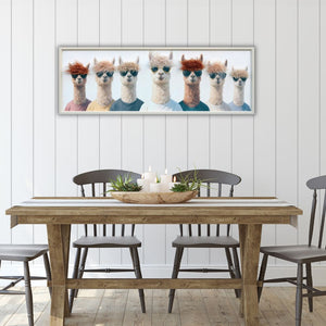 In einem hellen Esszimmer im dänischen Stil hängt eine Bild mit einem hellen Holzrahmen. Auf dem Bild sind sieben Lamas mit unterschiedlichen Frisuren aus Kunsthaar.