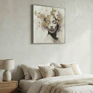 Ein Schlafzimmer mit einem quadratischen, gerahmten Gemälde einer Frau mit goldenen Blüten und Blättern im Haar, die über einem Bett mit beigen und cremefarbenen Kissen und Decken hängt. Eine Tischlampe auf einem Nachttisch aus Holz steht links davon. Die Wände sind in einem strukturierten, hellen Beigeton gehalten.