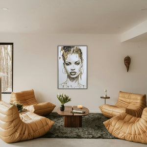 Luxuriöse Wohnzimmer-Lounge mit dem gerahmten XXL Frauenporträt-Gemälde „Gold covered Beauty“  an einer cremeweißen Wand. Das moderne Schwarz-Weiß-Kunstwerk mit goldener Metallfolie hängt mittig über einer gemütlichen Sitzecke bestehend aus ockerfarbenen Togo-Stil Ledersesseln, einem flauschigen grauen Teppich und einem organischen Holz-Couchtisch.