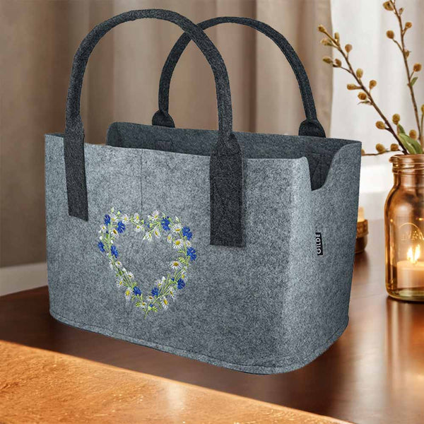 Auf einem Tisch steht eine Filztasche. Sie hat auf der Vorderseite ein Herz aus blauen und weißen Wildblumen.