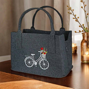Auf einem dunklen Holztisch steht ein grauer Filzshopper mit einem weißen eingestickten Fahrrad. Das fahrrad hat am Lenker einen Blumenkorb mit bunten Blumen.
