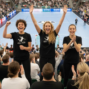 Drei jubelnde Damen stehen in einer Sporthalle inmitten von Fans. Sie tragen schwarze Handball Fan Damen T-Shirts, auf denen die Silhouette einer Handballspielerin und der Schriftzug „GRL PWR“ (Girl Power) in Weiß zu sehen sind. Die Frauen sind fröhlich, machen Siegergesten (erhobene Arme, Faust) und klatschen in die Hände, während sie ein Spiel auf dem blauen Spielfeld beobachten.