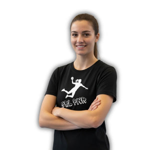 Eine lächelnde Frau mit braunem Pferdeschwanz trägt ein schwarzes T-Shirt. Auf dem Shirt ist eine weiße Silhouette einer Handballspielerin, die einen Wurf ausführt, abgebildet, darunter steht der Text "GRL PWR" (Girl Power). Die Frau steht mit verschränkten Armen vor einem weißen Hintergrund.