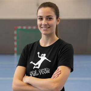 Nahaufnahme einer lächelnden Frau, die in einer Sporthalle steht. Sie trägt ein schwarzes Handball Fan Damen T-Shirt mit verschränkten Armen. Der weiße Druck zeigt eine weibliche Handball-Spielerin im Sprungwurf und den Slogan "GRL PWR" (Girl Power).