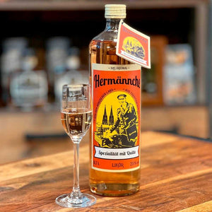 Eine Flasche Hermännche Quittenlikör  aus dem Bergischen Land mit einem markanten, orangefarbenen Etikett, das den Postboten Hermann und den Kölner Dom zeigt, steht auf einer Holztheke. Neben der Flasche Hermännche befindet sich ein gefülltes Likörglas. Im unscharfen Hintergrund sind weitere Flaschen in einer Destillerie zu erkennen.