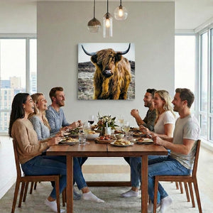 In einem hellen, modernen Esszimmer sitzt eine fröhliche Gruppe von Freunden an einem Holztisch beim gemeinsamen Essen, während an der weißen Wand im Hintergrund ein quadratischer Leinwanddruck eines schottischen Hochlandrindes hängt.