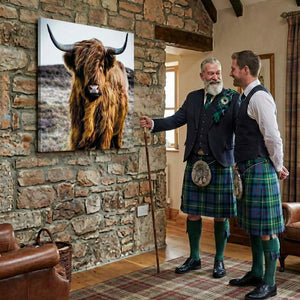 Zwei Männer in traditionellen Schottenröcken stehen in einem rustikalen Raum vor einer Steinwand und bewundern lächelnd einen großformatigen quadratischen Leinwanddruck eines braunen schottischen Hochlandrindes.