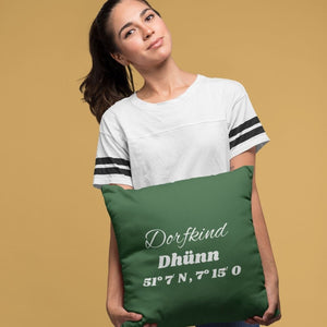 Eine frau hält ein grünes Kissen mit der weißen Bestickung Dorfkind Dhünn in den Händen. Das Kissen ist eine gute Geschenkidee für Dhünner aus der Region Bergisches Land.