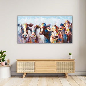 Breites Wandbild mit fünf bunten, abstrakt gemalten Kühen, das über einem hellen Holz-Sideboard mit Rattan-Details an einer grauen Wand hängt. Die Tiere blicken frontal nach vorne und leuchten in kräftigen Farben. Der Raum ist hell gestaltet, mit Holzboden und dezenter Pflanzendeko auf einem Beistelltisch.