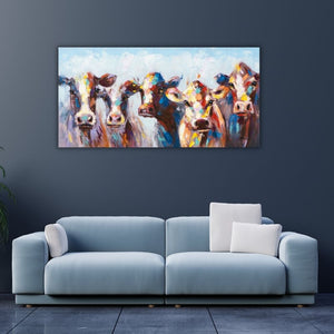 Ein breites, farbenfrohes Gemälde mit fünf stilisierten Kühen hängt an einer dunkelblauen Wand über einem modernen, hellblauen Sofa mit weißen Kissen. Die Tiere sind in kräftigen Tönen wie Orange, Gelb, Rot und Blau mit sichtbaren Pinselstrichen gemalt und blicken den Betrachter direkt an. Der Raum wirkt modern und kühl, ergänzt durch einen dunklen Fliesenboden und eine angeschnittene Pflanze am linken Bildrand.