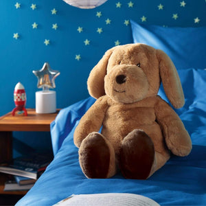 Kuschelset Dream Team Hund Max sitzt auf dem Bett. Das Stofftier von Biederlack hat im Innern eine weiche Schmusedecke.