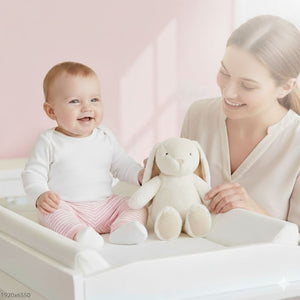 Ein Baby sitzt lachend auf dem Wickeltisch mit dem Stofftier Hase Nelly in der Hand.