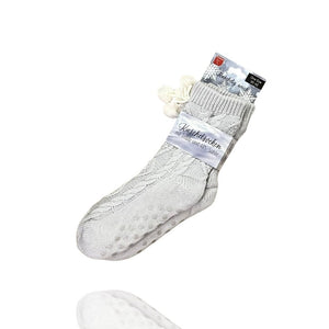 Nahaufnahme einer einzelnen, grauen Strick-Socke/Hausschuh oder Kuschelsocken. Die Socke hat ein ausgeprägtes Zopfmuster und ein weiches, cremefarbenes Plüschfutter am oberen Schaft, verziert mit dekorativen Pompons. Die Unterseite zeigt Anti-Rutsch-Noppen.