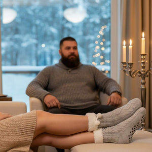 In einem Wohnzimmer hat eine Frau graue wärmende Kuschelsocken an Sie sitzt auf dem Sofa und hinter ihr sitzt ihr Mann.