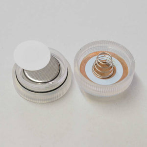 Dieses Bild zeigt zwei separate Teile eines kleinen, runden LED-Lichts. Auf der linken Seite ist die Basis des Lichts zu sehen, die eine Knopfzelle mit einer runden, weißen Isolierscheibe darüber enthält. Der obere Teil des Sockels ist transparent und geriffelt. Auf der rechten Seite ist die passende obere Kappe des Lichts zu sehen, die ebenfalls transparent und geriffelt ist und eine kleine Metallfeder enthält, die dazu dient, die Batterie zu kontaktieren. 