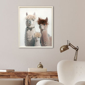 An einer Wand hängt eine gerahmtes Bild von drei lamas. Jedes lama hat durch das Kunstfell eine eigene Frisur. das Bild heisst Stylish Lama Family und hängt über einem Tisch.