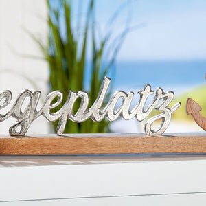 Der Scgriftzug Liegeplatz mit Anker steht auf einem Sideboard. Dieses maritime Dekoelement spricht alle Nord- und Ostseefans an aber auch alle Yacht und Bootsbesitzer.e