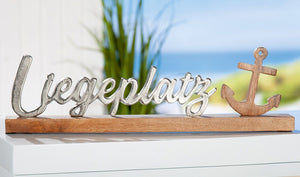 Der Scgriftzug Liegeplatz mit Anker steht auf einem Sideboard. Dieses maritime Dekoelement spricht alle Nord- und Ostseefans an aber auch alle Yacht und Bootsbesitzer.e