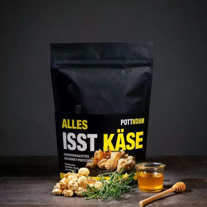 Alles isst Käse steht in gelb weißen großen Buchstaben auf einer schicken schwarzen Tüte. Vor der Tüte liegt Popcorn, Oregano und ein Glas mit Honig.