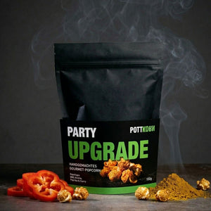 Eine schwarze, matte Standbeuteltüte der Marke „POTTKORN“ steht im Zentrum vor einem dunklen Hintergrund. Die Banderole trägt den großen grünen Schriftzug „PARTY UPGRADE“ und den Untertitel „Handgemachtes Gourmet-Popcorn“. Links neben der Tüte liegen frische, rote Paprikaringe, rechts ein kleiner Haufen gelbes Currypulver. Einzelne goldbraun karamellisierte Popcorn-Stücke sind um die Zutaten verteilt. Hinter der Tüte steigen feine, graue Rauchschwaden auf, die eine würzige, rauchige Atmosphäre (BBQ-Aroma) s