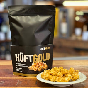 Auf einer Theke steht eine schwarze Tüte. Auf ihr steht Hüftgold von Pottkorn. Handgemachtes Butterkaramell Gourmet Popkorn. Davor steht ein kleiner Teller mit Popkorn aus Mushroom Mais.