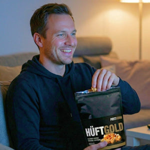 Ein Mann sitzt auf einer Couch und isst aus einer schwarzen Tüte das Butterkaramell Popcorn mit dem Namen Hüftgold von Pottkorn.